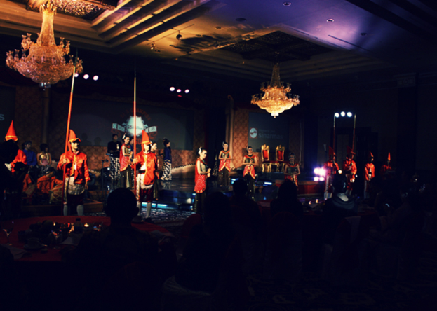 Eine Gruppe von Menschen sitzt und schaut eine Sendung mit zwei Kronleuchtern an der Decke.