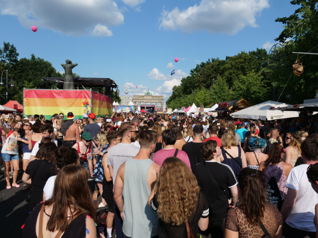 Eine große Menschenmenge geht eine Straße mit Zelten, Bäumen, Pfählen, Lichtern und einer Statue entlang während des Christopher Street Day Festivals in Berlin, mit Gebäuden und einem Himmel voller Wolken und Ballons im Hintergrund.