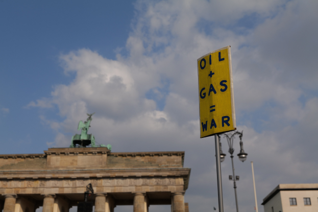 Das Brandenburgische Tor in Berlin, Deutschland, mit einem gelben Schild, auf dem "Krieg um Öl und Gas" steht, im Vordergrund vor einem bewölkten Himmel.
