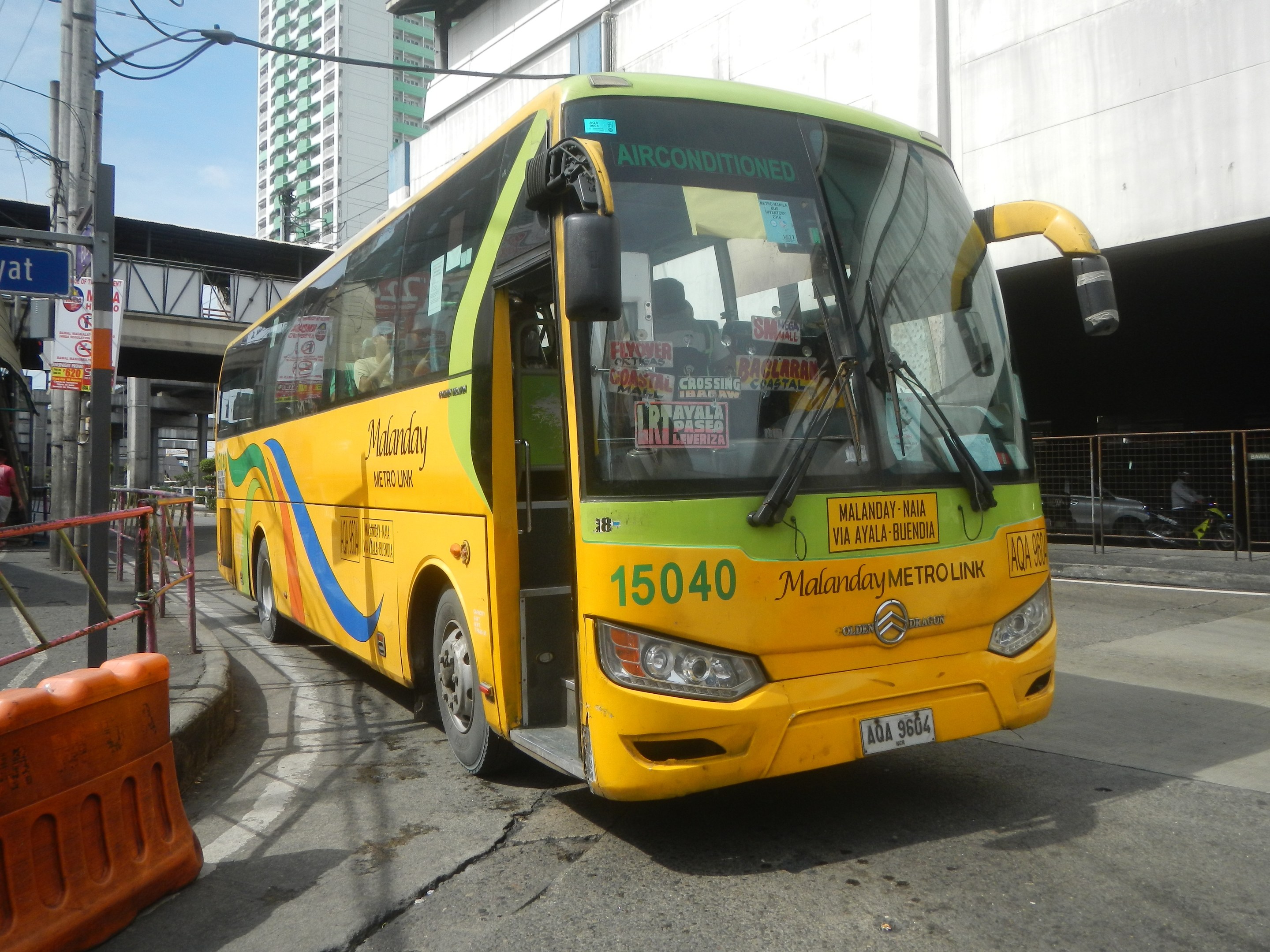 Ein gelber Bus fährt auf einer Straße mit hohen Gebäuden, einem Geländer auf der linken Seite, Fußgängern auf dem Gehweg und einer Brücke im Hintergrund unter dem Himmel.