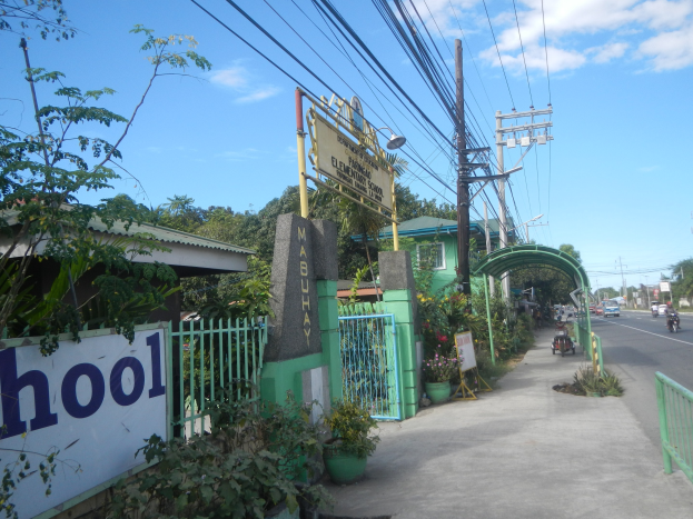 Schulwegweiser am Straßenrand mit einem Torbogen, einem Tor und einer Namenschilder, umgeben von Fahrzeugen, Pflanzen, Bäumen, Strommasten und einem bewölkten Himmel.