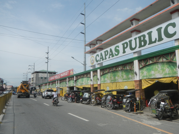 Eine belebte Stadtstraße mit Fahrzeugen, einem Geländer auf der linken Seite, Gebäuden, Pfählen, Laternen, Schildern und einigen Menschen unter einem bewölkten Himmel, mit einem Gebäude mit der Aufschrift "Capas Public".