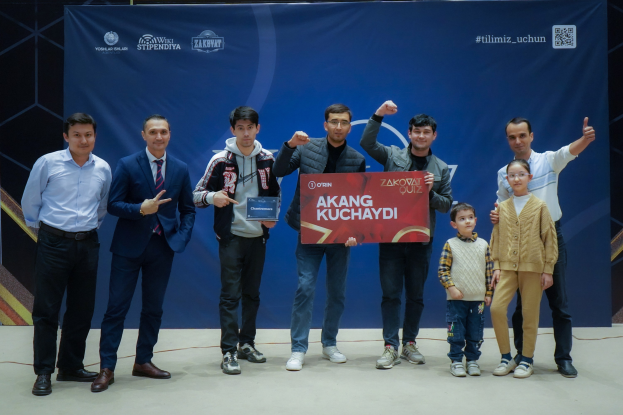 Gruppe von Menschen auf einer Bühne, die eine Tafel mit dem Text "Akang Kuchaydi - Zakovat Quiz" halten, mit einem Banner und einem Strichcode im Hintergrund.