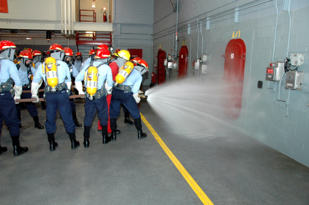 Feuerwehrleute in Helmen und Ausrüstung spritzen sich gegenseitig mit Wasser an, mit Equipment wie Rohren, Feuerlöschern und einer Leiter im Hintergrund.