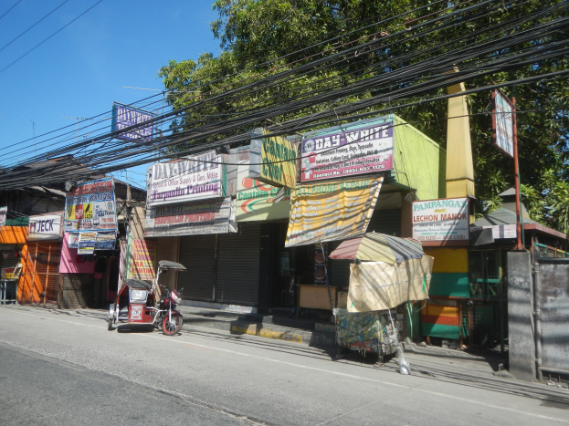 Eine Straßenszene mit einem geparkten Motorrad, Gebäuden, Geschäften, Namensschildern, Strommasten, Stromkabeln, Bäumen und einem klaren blauen Himmel.