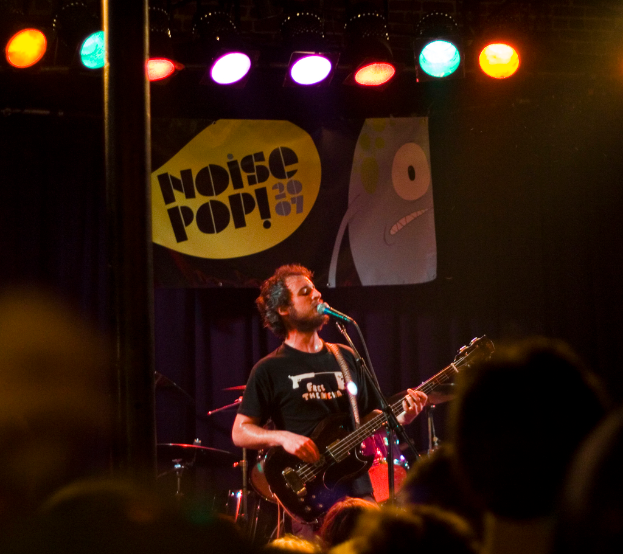 Eine Person in einem schwarzen Kleid singt und spielt Gitarre auf der Bühne, mit Trommeln und einem blauen Vorhang mit einem Plakat dahinter und einer Menge davor.