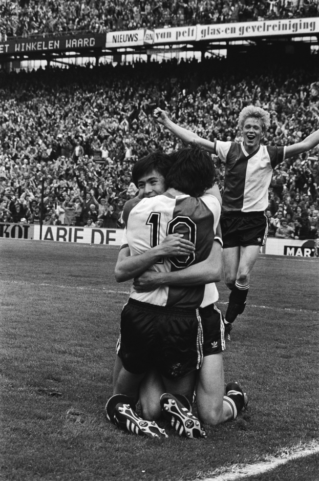 Drei Fußballspieler feiern nach einem Tor auf dem Boden, mit jubelnder Menge und schwenkenden Fahnen im Hintergrund, in einem schwarz-weißen Bild.