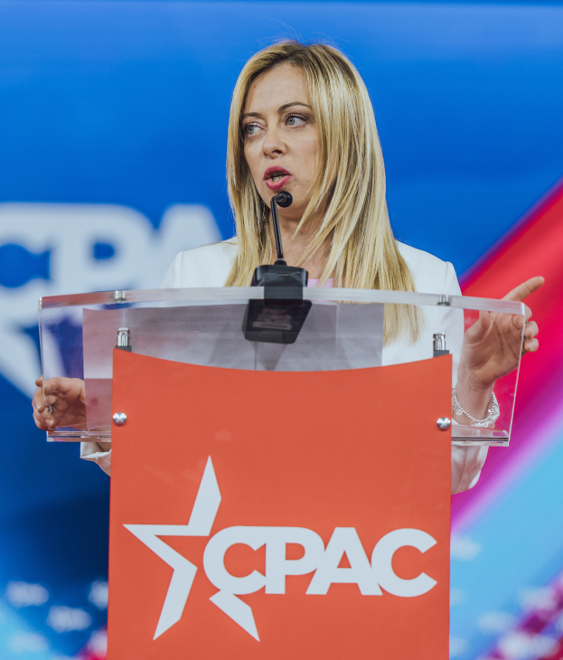 Eine Frau in einem weißen Kleid steht an einem Rednerpult mit einem Mikrofon, wobei sie wie eine Politologin wirkt, mit einem unscharfen Hintergrund.