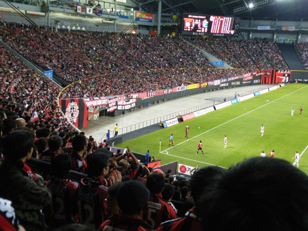 Ein Fußballspiel in einem großen Stadion mit Spielern auf dem Feld und Zuschauern in den Rängen, mit Bannern, einem Zaun, einem Display, Metallstangen und Deckenleuchten.