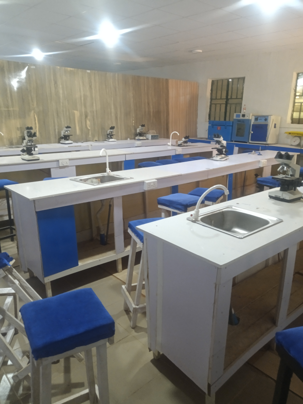 Ein Labor mit blauen Hockern und weißen Arbeitsplatten, ausgestattet mit einem Waschbecken mit Wasserhähnen, verschiedenen Maschinen, einer Wand mit Fenstern und Deckenleuchten, Teil der Universität für Wissenschaft und Technologie in Ahmedabad.