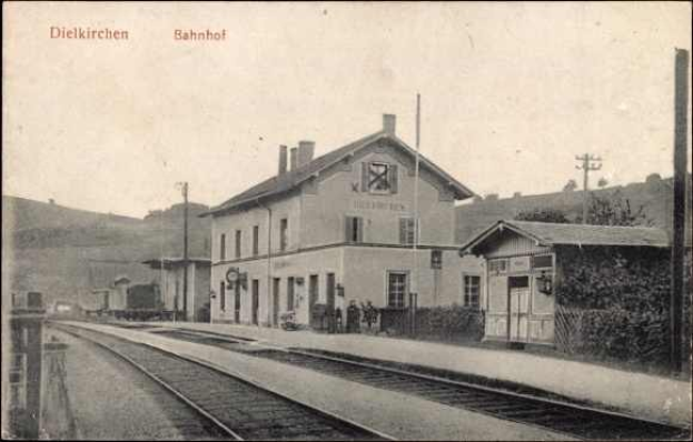 Altes Schwarz-Weiß-Foto des Bahnhofs Dielkirchen in Deutschland mit einem Zug auf den Schienen, umgeben von Häusern, Strommasten, Bäumen und Menschen, mit Hügeln und einem klaren Himmel im Hintergrund.