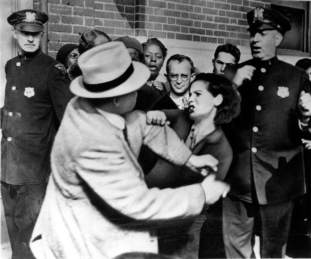 Zwei Personen kämpfen in einem Schwarz-Weiß-Bild, umgeben von mehreren anderen, einige tragen Mützen, und eine Wand im Hintergrund.