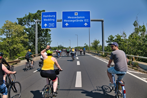 Eine Gruppe von Radfahrern mit Helmen fährt eine Straße mit einer Begrenzung auf einer Seite und Bäumen auf der anderen entlang, unter einem klaren blauen Himmel mit Laternen im Hintergrund; ein Schild oben zeigt eine Radtour in Hamburg.