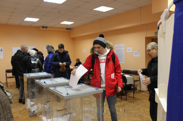 Eine Gruppe von Menschen steht um eine Wahlurne in einer Wahllokal, einige halten Papiere, mit einem blauen Vorhang und weißem Objekt auf der rechten Seite, Stühle, Tische, Papiere an der Wand und Deckenlampen im Hintergrund.