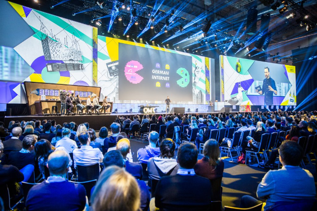 Eine große Menschenmenge sitzt auf Stühlen vor einer Bühne, einige Personen stehen im Vordergrund auf dem Podium, ein Tisch mit verschiedenen Gegenständen links und Bildschirme und Lichter im Hintergrund auf der Deutschen Internet-Konferenz 2019.