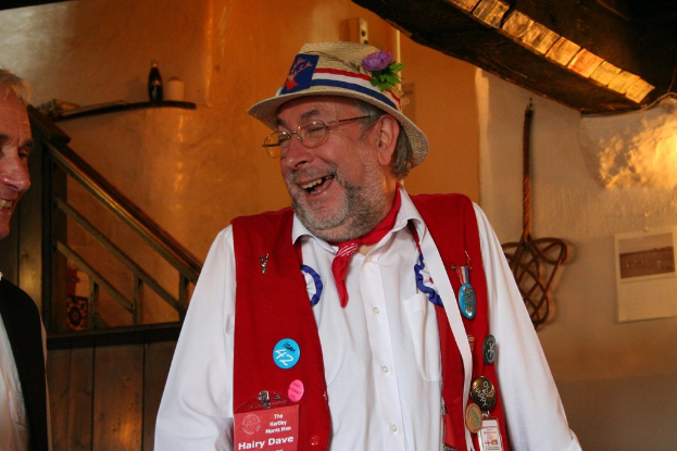 Ein Mann in einem weißen Kleid, Hut und Brille lächelt, während er neben einem anderen Mann in einem Blazer steht; im Hintergrund ist ein hölzerner Stock an der Wand neben einem Kalender angebracht, mit einer Flasche und anderen Gegenständen auf einem hölzernen Regal darüber.