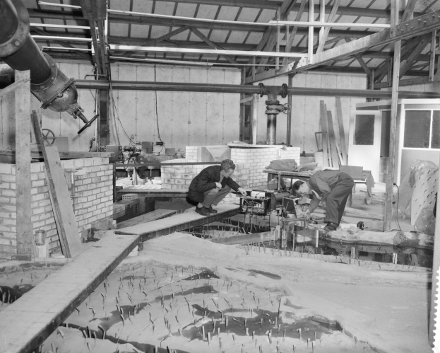 Zwei Männer arbeiten an einem Holzstück in einer schwarz-weißen Fabrikumgebung mit Maschinen, Rohren und Fenstern.