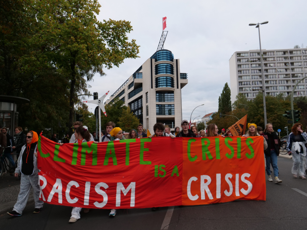 Eine Gruppe von Menschen marschiert eine baumbestandene Straße entlang und hält ein Banner mit der Aufschrift "Klima-Krise ist eine Krise", mit Gebäuden und einem klaren Himmel im Hintergrund.