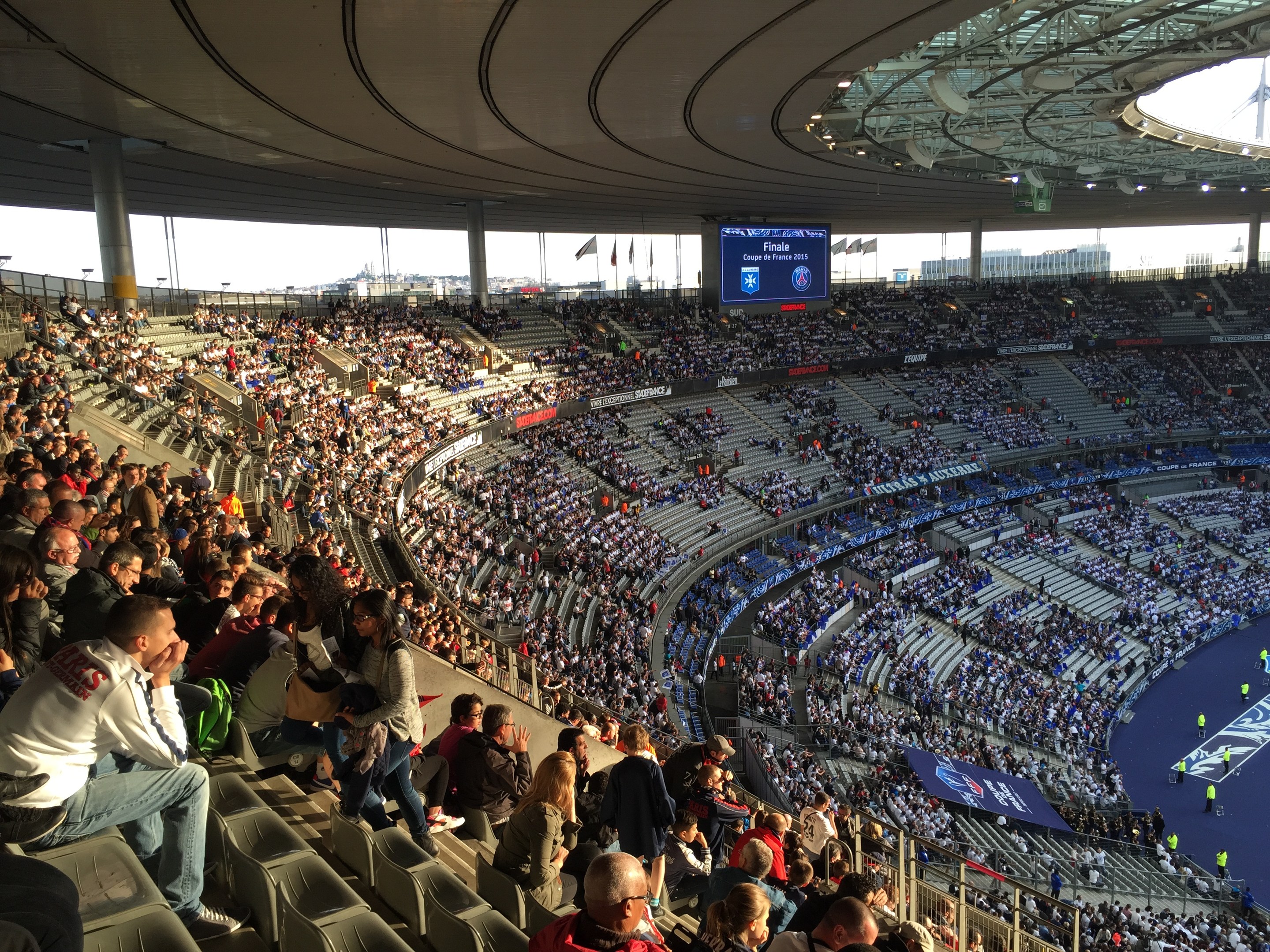 Große Zuschauermenge in einem Stadion bei einem Fußballspiel mit einer Bühne rechts, Fahnen, Stangen, einem Bildschirm und der Allianz Arena in München, Deutschland im Hintergrund.