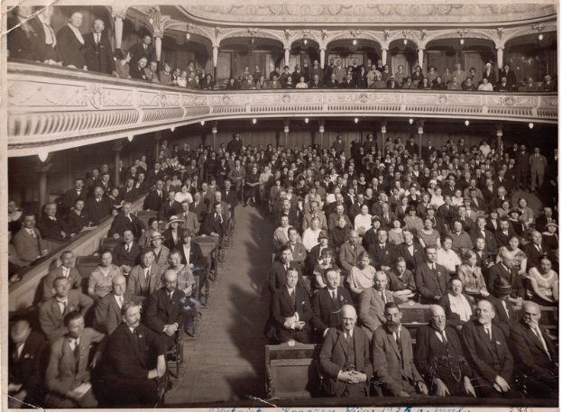 Eine große Gruppe von Menschen, einschließlich Männer und Frauen, die in einem Theater sitzen, in einem alten Schwarz-Weiß-Foto abgebildet.