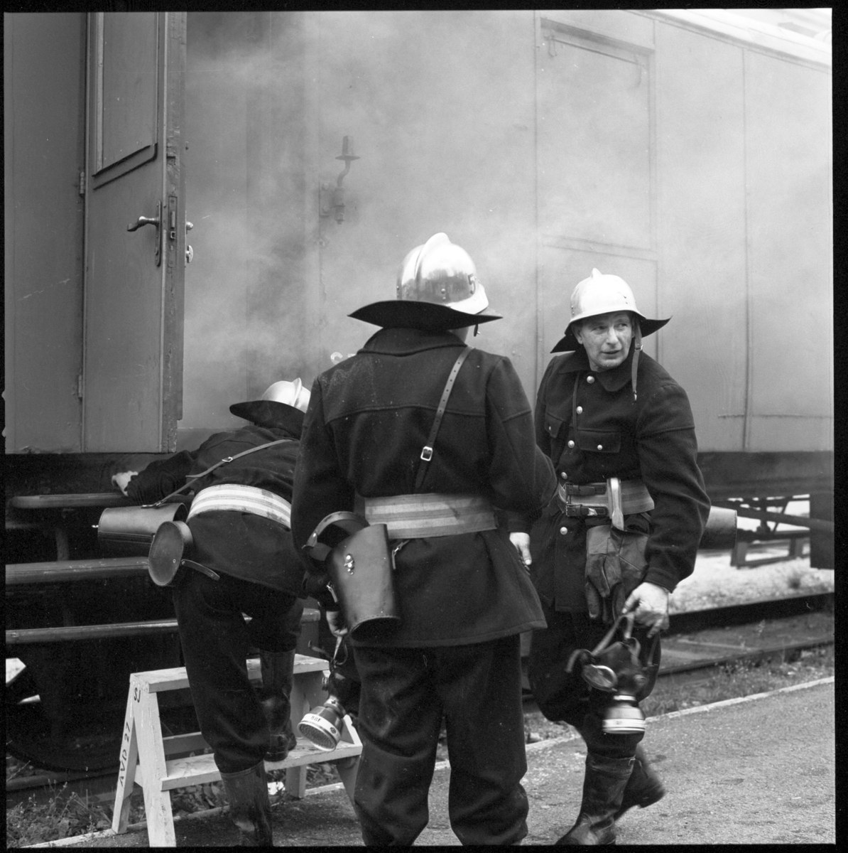 Feuerwehrmänner in Helmen stehen auf einem Bahngleis mit einem auf einem Hocker sitzend, während ein Zug im Hintergrund Rauch ausstößt in einem Schwarz-Weiß-Bild.