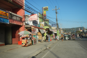 Eine belebte Stadtstraße mit Menschen, umgeben von Gebäuden, Geschäften, Schildern, Polen, Drähten, Bäumen und Hügeln unter einem klaren blauen Himmel.