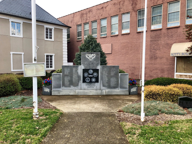 Ein Denkmal für Opfer des Zweiten Weltkriegs steht vor einem Backsteingebäude mit einem Schild, umgeben von Grün und Pfählen, mit einem Himmel im Hintergrund.