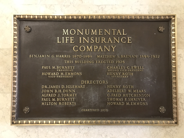 Plakette an einer Gebäudewand mit der Aufschrift "Monumental Lebensversicherungsgesellschaft" in fetter Schrift und einem dekorativen Rahmen, auf dunklem Material montiert.