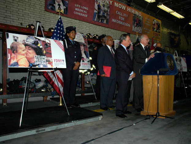 Eine Gruppe von Menschen in der Nähe eines Podiums mit Mikrofonen, Schildern auf Ständern, Flaggen und Schildern an einer Wand, alles unter hellen Scheinwerfern.