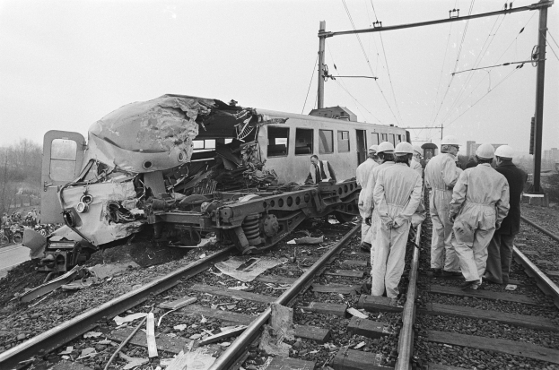 Schwarz-weiß-Bild einer Zugkollision auf den Schienen mit Einsatzkräften und beschädigter Infrastruktur im Hintergrund.